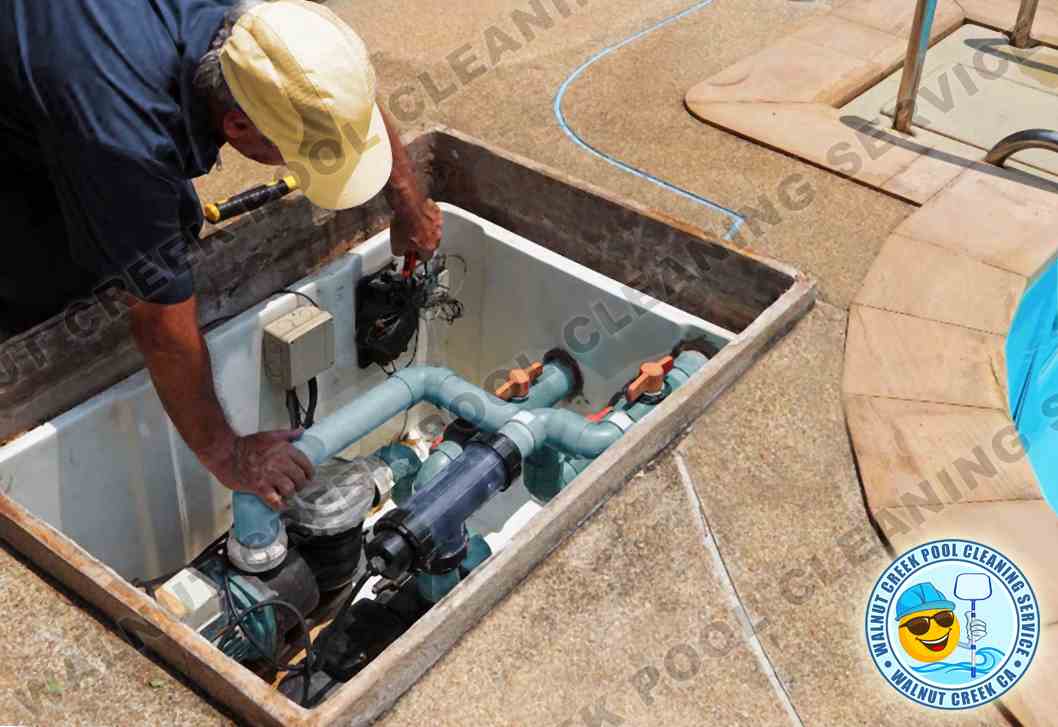 A pool service professional from Walnut Creek Pool Cleaning Service works on pool equipment within an open underground pit. The technician, wearing a yellow cap and blue shirt, is leaning over the pit and appears to be repairing or maintaining the pool's plumbing system, which includes pipes, valves, and pumps. The surrounding area is paved with light-brown tiles, and a blue pool hose stretches across the scene, indicating active maintenance work.