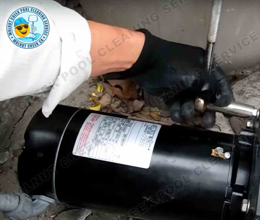 A Walnut Creek Pool Cleaning Service technician is repairing a pool pump, with the focus on their hands wearing black gloves, using a wrench on the pump's metallic components. The label on the pump is visible, indicating technical details, and the environment suggests outdoor ground with scattered leaves, indicating an on-site repair service.